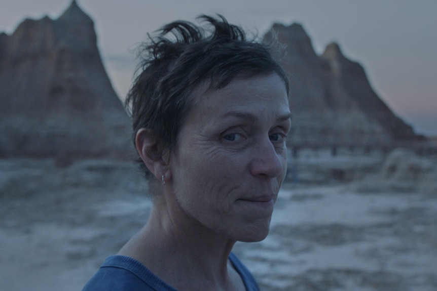 Twilight shot of the actress with pixie haircut and no make-up, wearing blue t-shirt, with arid mountain landscape in background