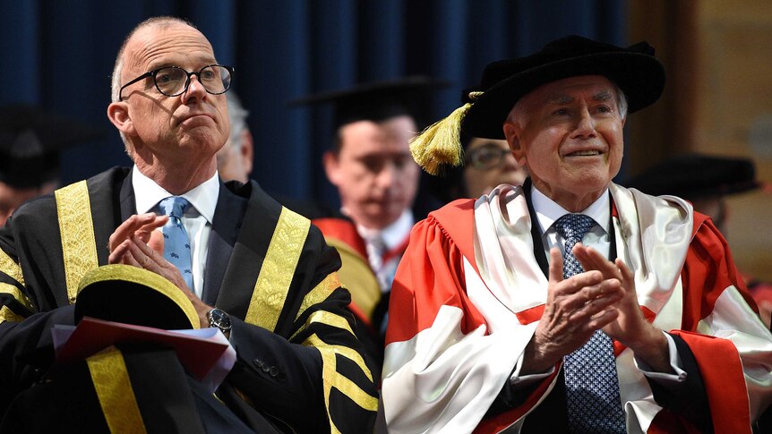 John Howard, wearing gown and mortarboard, sits next to University of Sydney Vice Chancellor Michael Spence