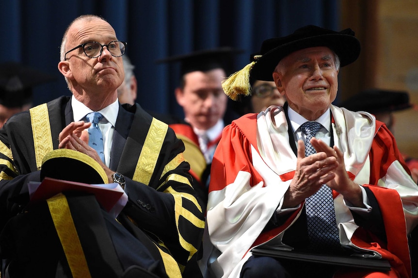 John Howard, wearing gown and mortarboard, sits next to University of Sydney Vice Chancellor Michael Spence