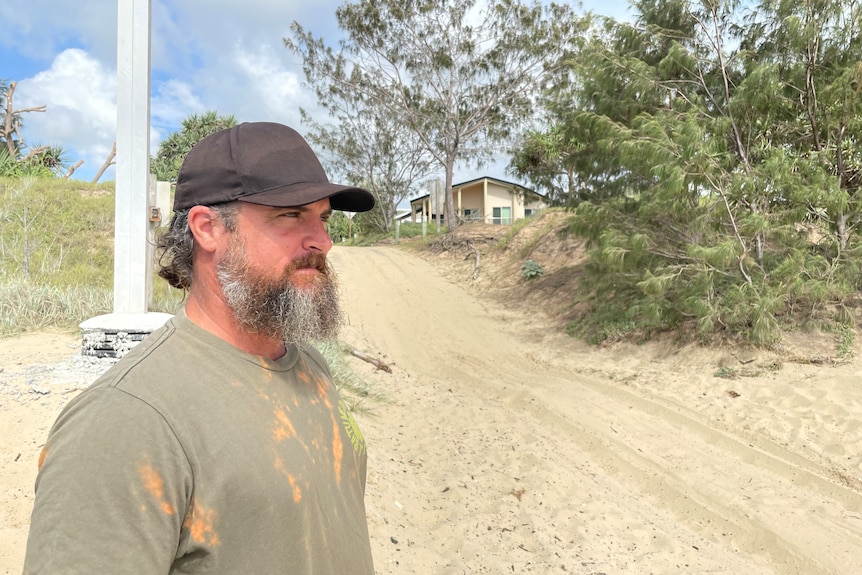 A man with a black cap on stands in front of a vehicle access point to a beach