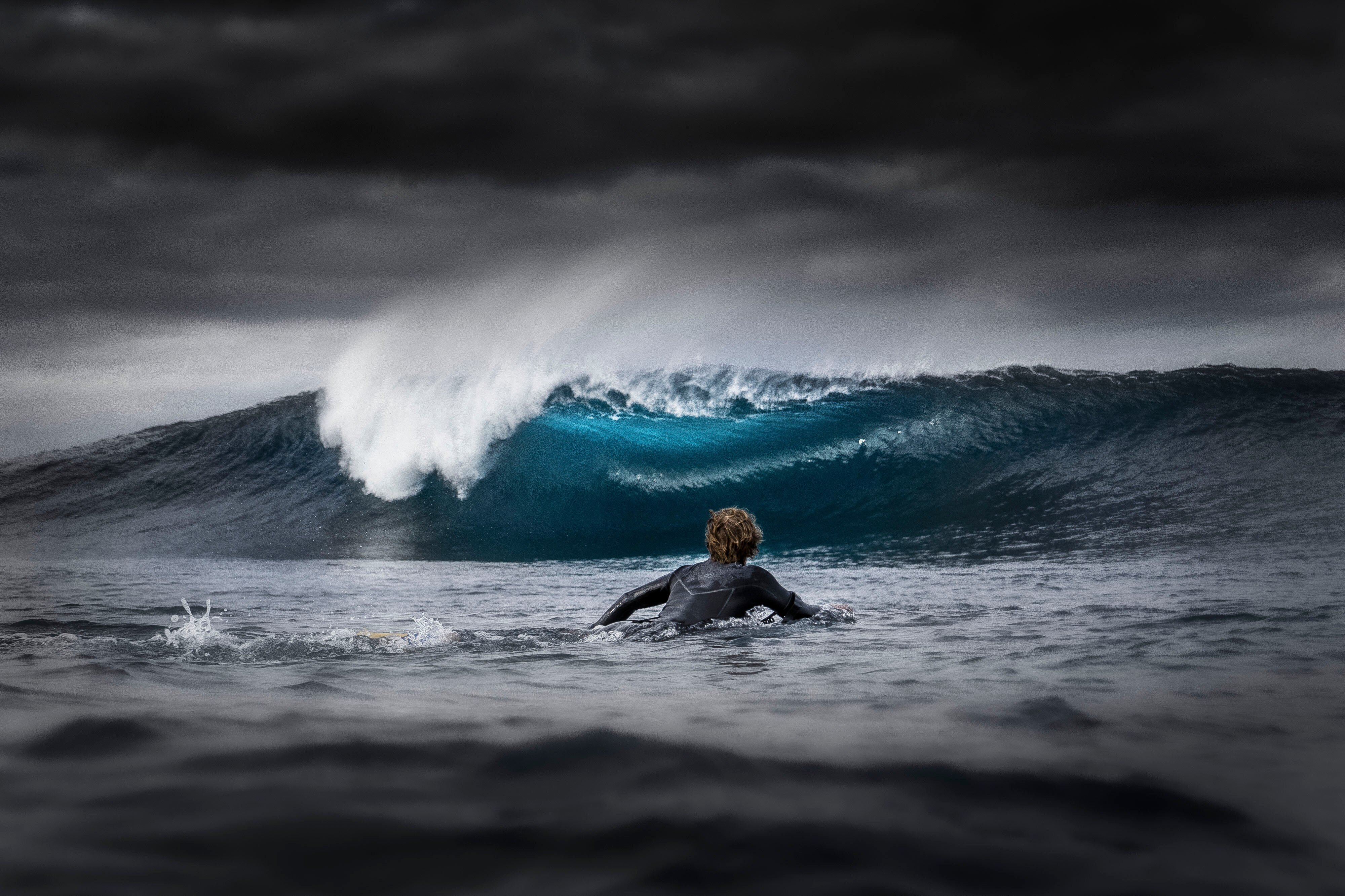 A bodyboarder paddles out to sea, while big storm clouds block the sun and create an electric blue colour effect on the wave