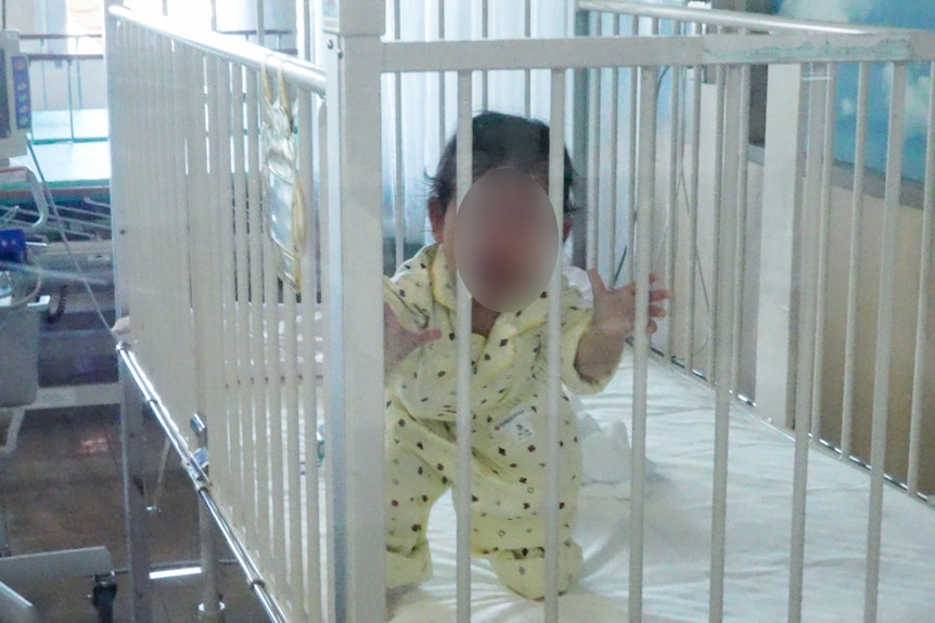 A little baby girl in yellow pyjamas grasping at the bars of her cot in hospital 