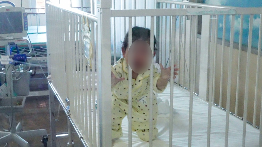 A little baby girl in yellow pyjamas grasping at the bars of her cot in hospital 