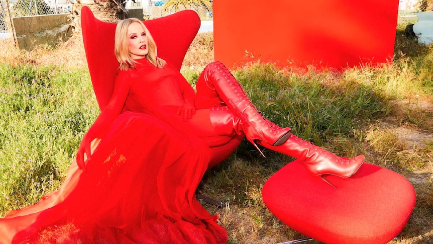 Kylie Minogue lounges in a red chair and foot stool in matching red catsuit outdoors on an unkempt lawn
