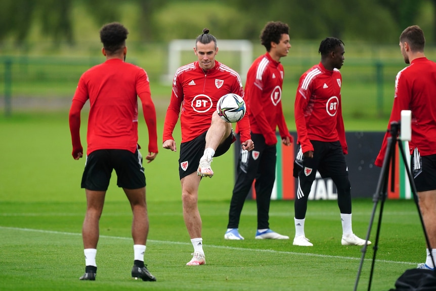 Star Welsh footballer Gareth Bale juggles a ball on his knee as his teammates stand around at training.