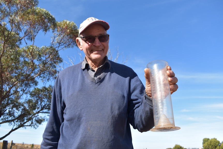 An older, fair-skinned man, Lloyd, smiles in transition lenses and ratty cap as he holds up an empty rain gauge.