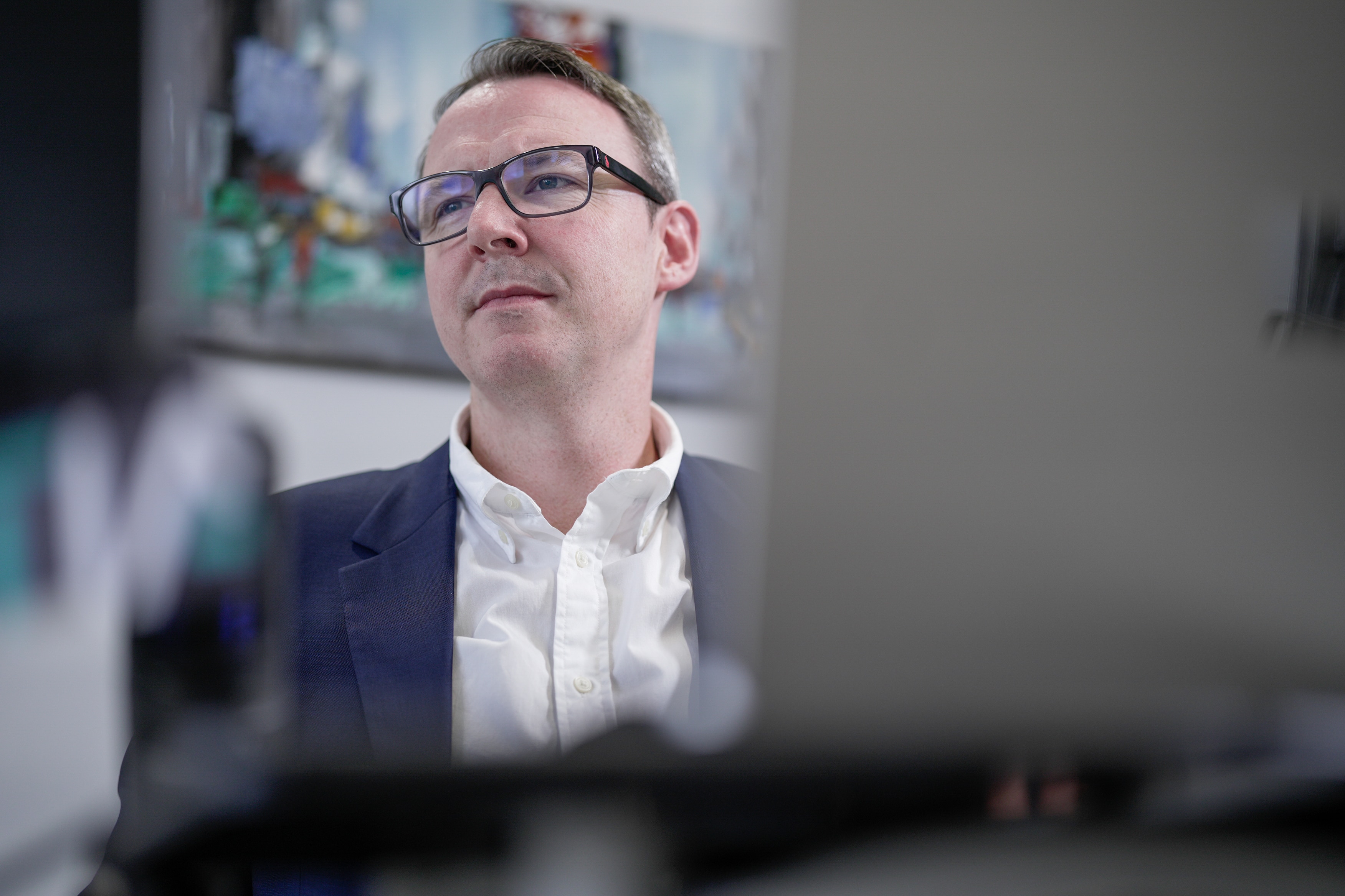 A man in a blue suit and glasses sits at a desk with a laptop in foreground.