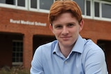 A smiling young man with ginger hair stands in front of a brick building bearing the lettering "Rural Medical School".