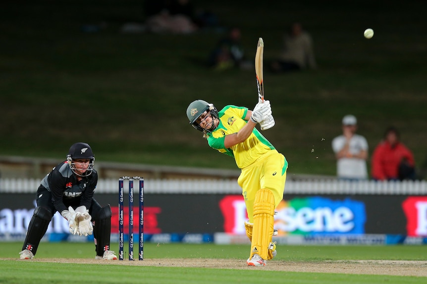 An Australian women's cricketer hits a lofted drive down the ground as the wicketkeeper watches behind the stumps.