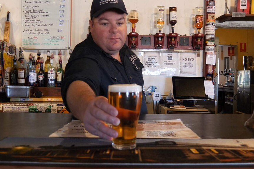 A man in a dark T-shirt and cap leans over a hotel bar as he serves a frothy beer