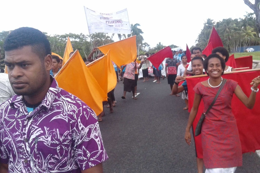 A protest in Fiji. People wave yellow and red flags as they walk down a road.