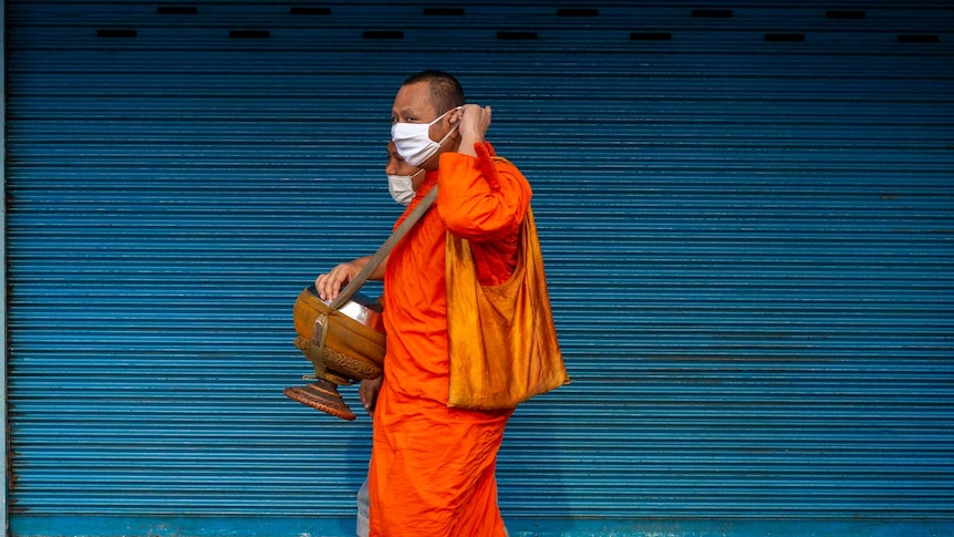 A Buddhist monk in Thailand in a face mask walking past a bright blue wall