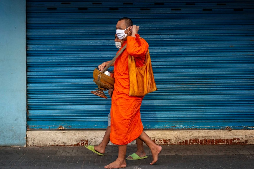 A Buddhist monk in Thailand in a face mask walking past a bright blue wall