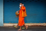 A Buddhist monk in Thailand in a face mask walking past a bright blue wall