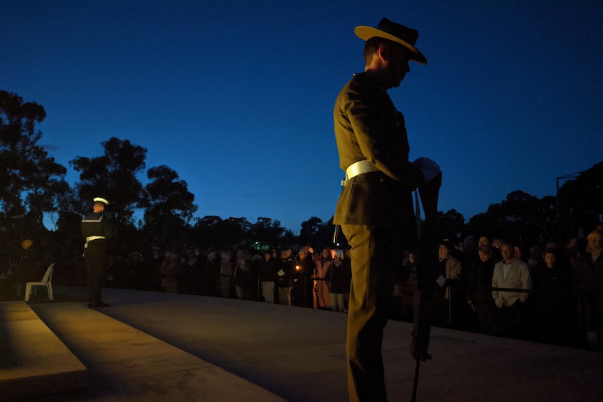 A soldier stands with his head bowed at a dawn service with light turning blue behind him.