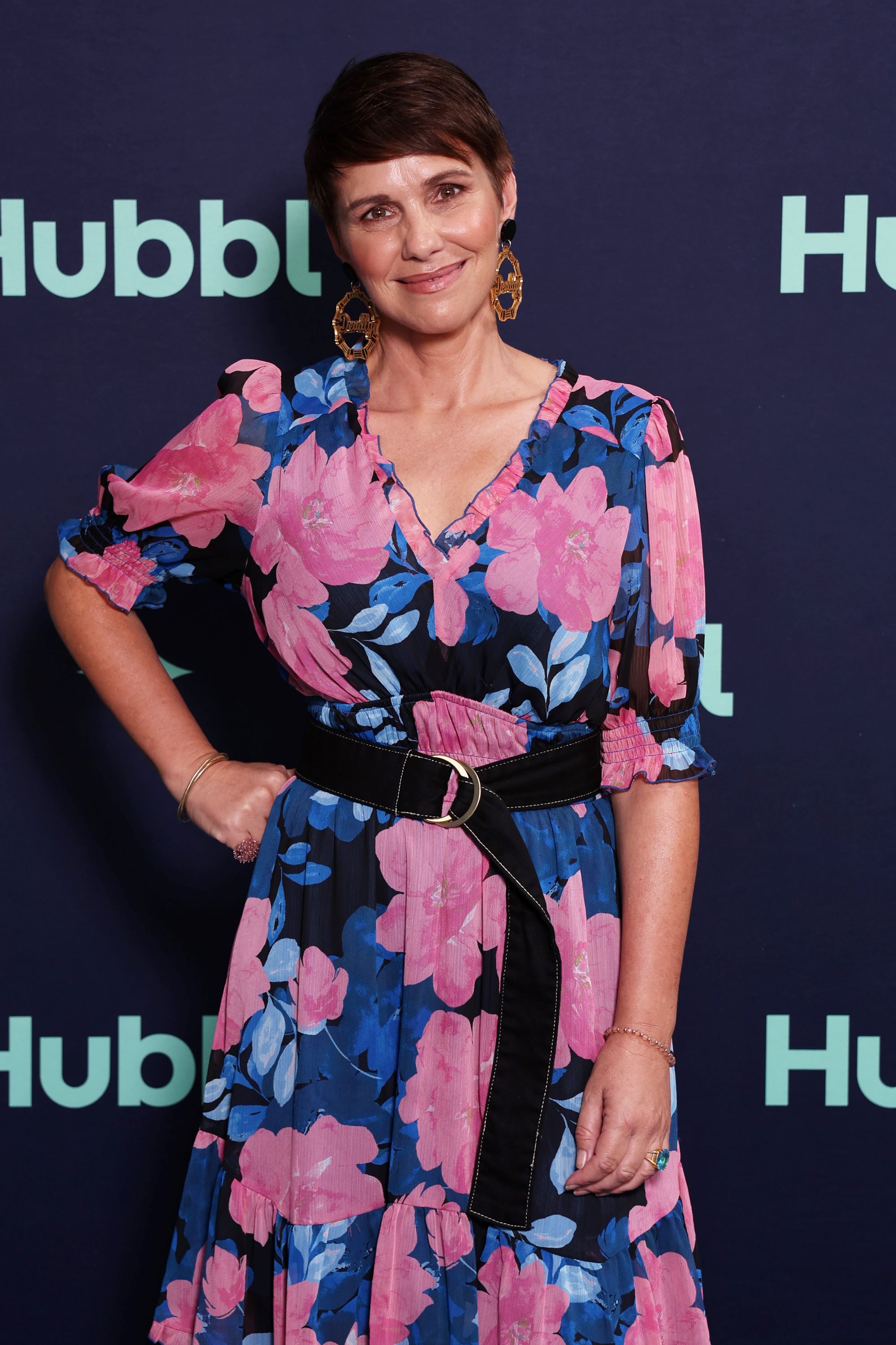 A woman with short brown hair and a wide smile wearing a pink and blur floral dress, standing with her hand on her hip