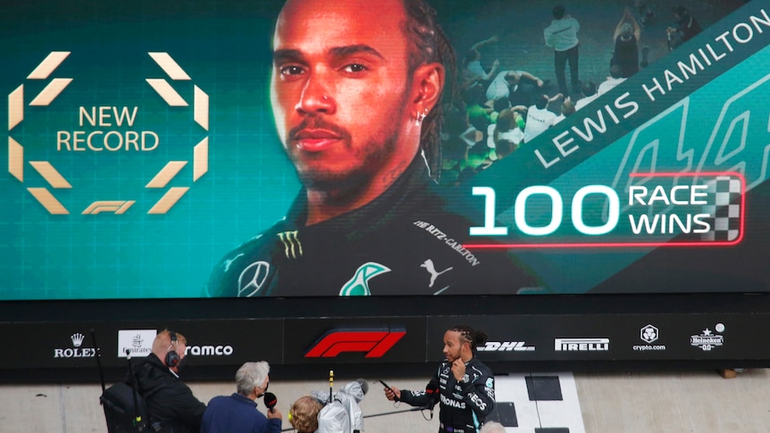 Lewis Hamilton is interviewed by a TV crew in front of a 100 race wins billboard
