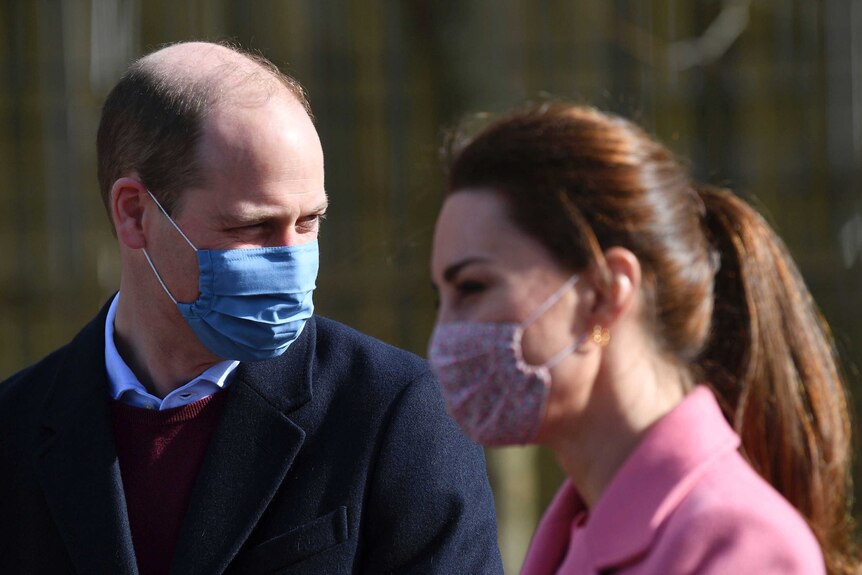 Prince William wearing a medical mask looks behind him. Blurred in the foreground, Kate looks ahead, also wearing a mask.