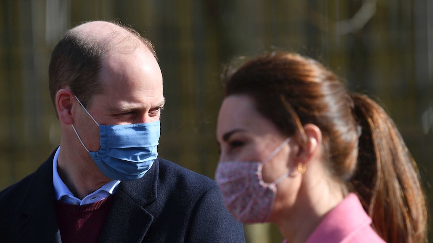 威廉王子戴着医用口罩看着他身后。 前面不显眼的凯特看着前方，也戴着口罩。