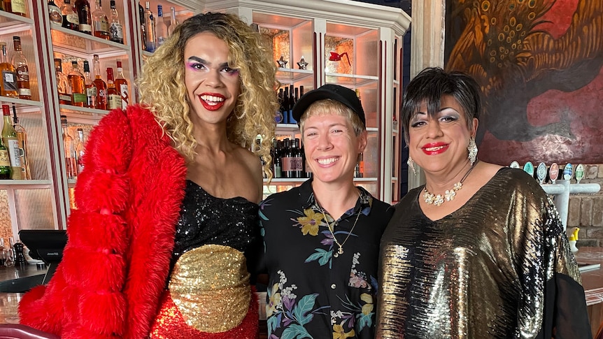 Mon Schafter standing between two Indigenous draq queens in costume in a bar.