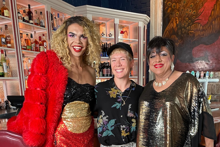 Mon Schafter standing between two Indigenous draq queens in costume in a bar.