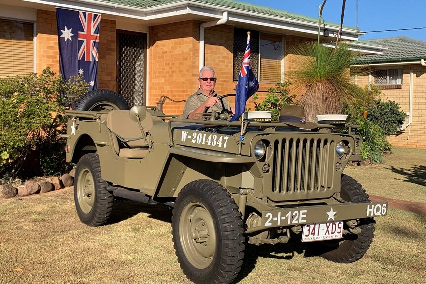 A man sits in an Army jeep parked in the front yard of a house