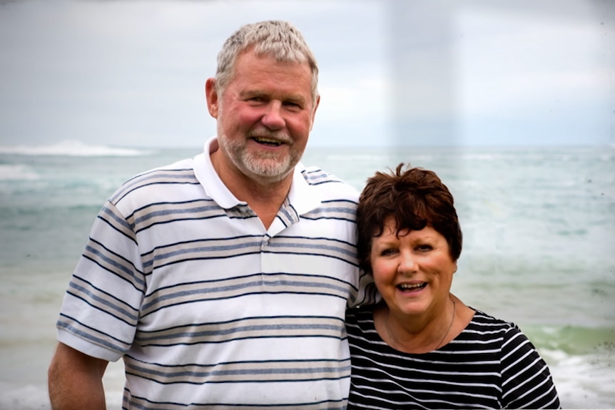 A husband and wife stand together smiling in front of the ocean.