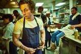 A young white man in a white tshirt and blue apron, stands in a busy commercial kitchen looking stressed 