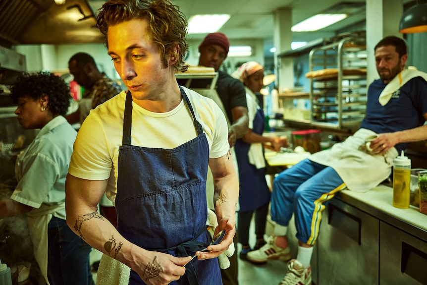 A young white man in a white tshirt and blue apron, stands in a busy commercial kitchen looking stressed 