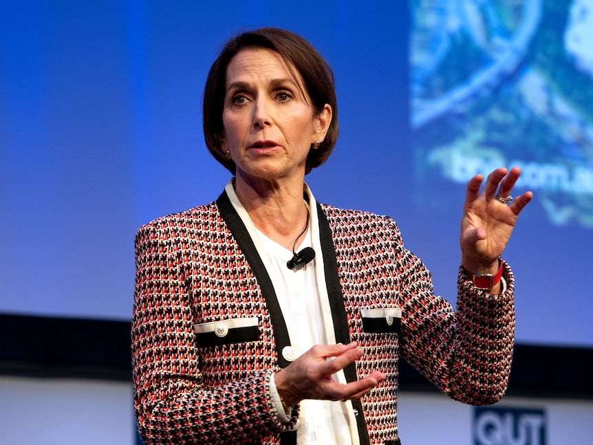 A woman with short brown hair wearing a black and red patterned blazer talking on stage in front of a blue background
