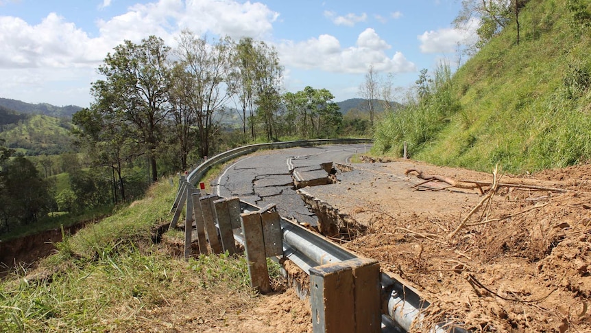 A downhill sloping road is covered in dirt and has deep cracks in it. On one side the barriers are twisted and warped.