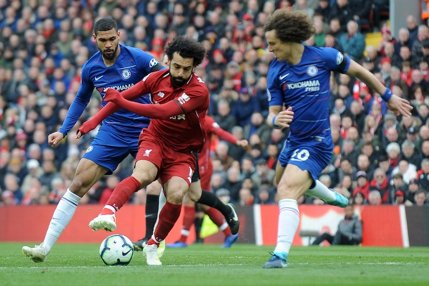 A man reaches for a soccer ball with his right foot as two opponents close in on him