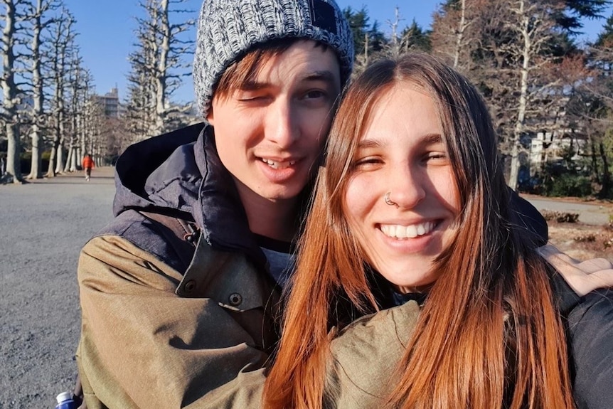 A young man and woman wearning winter clothes embrace as they smile at the camera in winter park setting