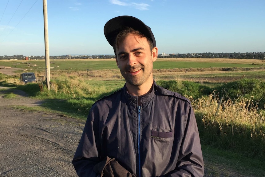 A man standing in a field holding a phone, smiling at the camera.