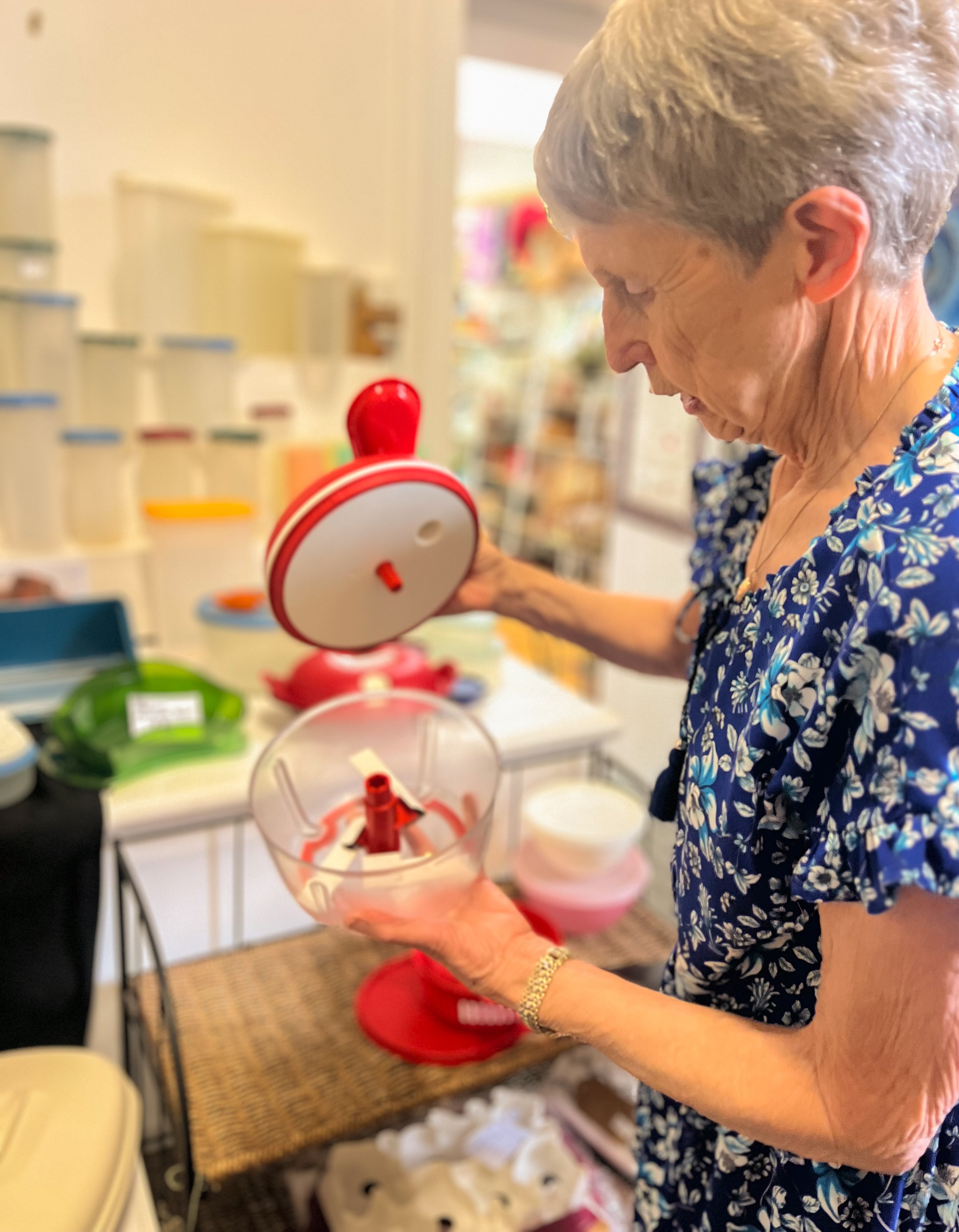 Uma senhora vestida de azul olha para um tupperware vermelho na frente de uma loja
