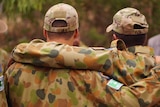 Two teenage boys photographed from behind in camouflage army uniforms with their arms around each other's shoulders.