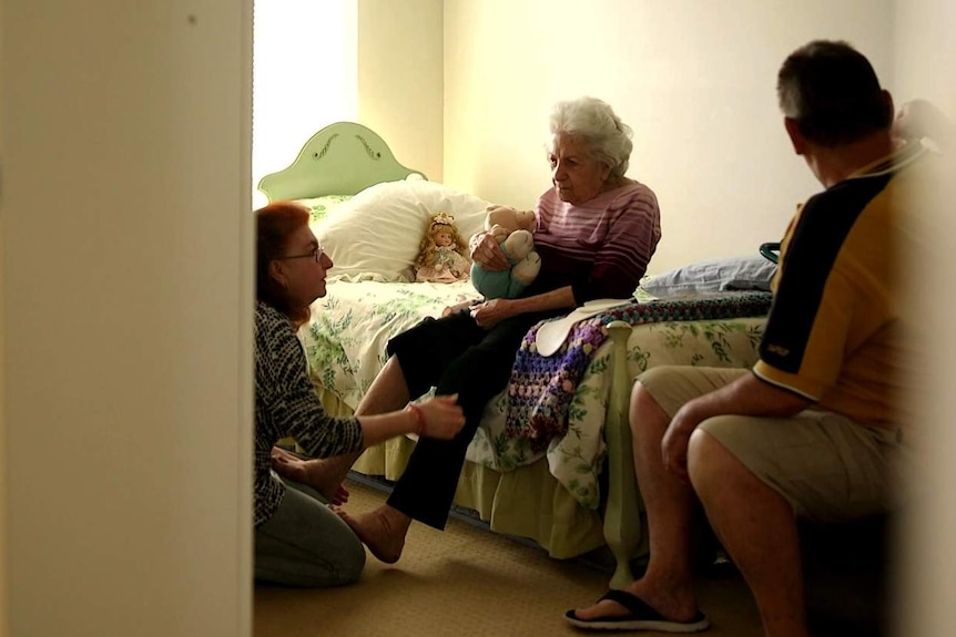 An elderly woman sits on a bed while a younger woman kneels on the floor helping her.