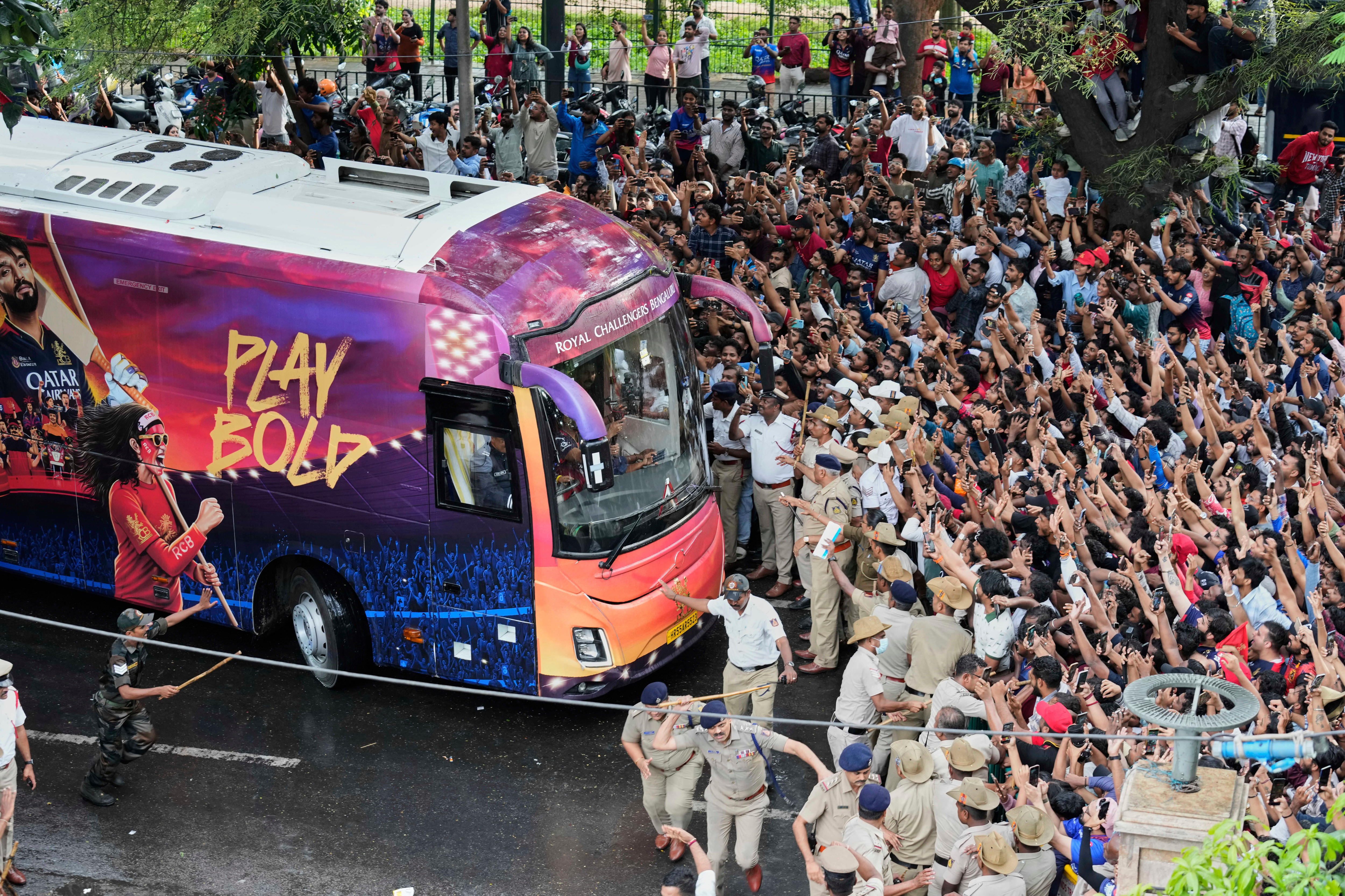 A purple and blue bus with 'Play Bold' on the side approaching a large crowd of fans with their arms in the air