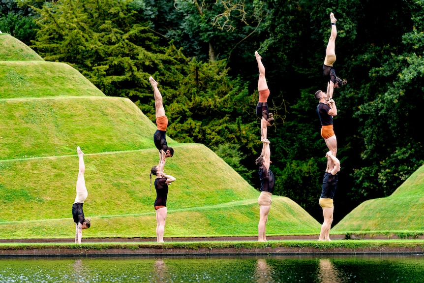 Eight circus artists perform handstands, some stacked two and three high, in front of a grassy terraced slope 