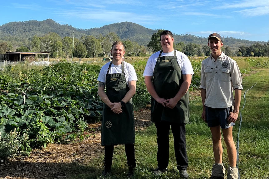 Three people stand smiling next to a garden of vegetables and mountains in the background.