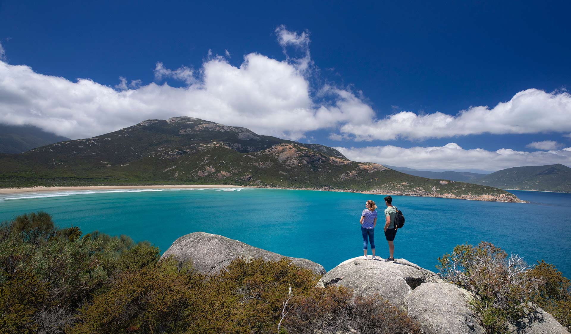Mountains surround an ocean bay in Wilsons Promontory National Park. Two hikers stand on a cliff.