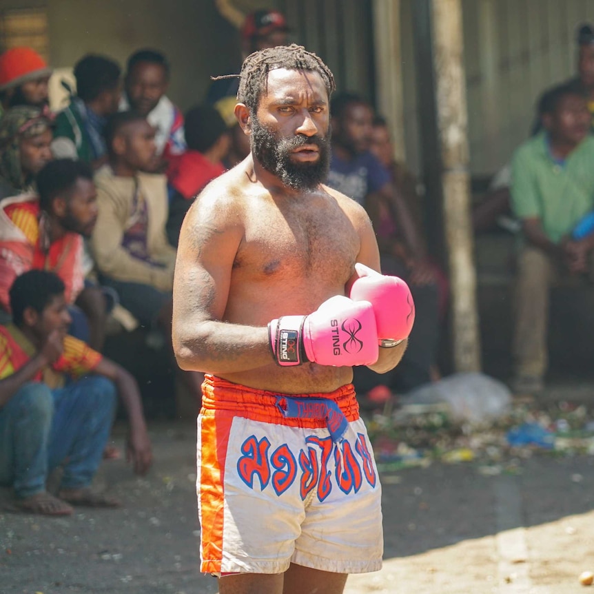 A man in boxing gloves and shorts stands in an arena as others look on