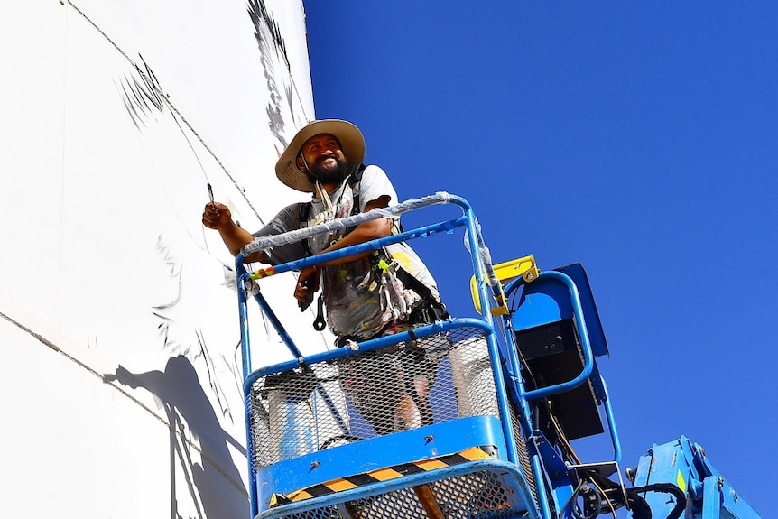 A man is leaning on a cage in the air, smiling. He has a paintbrush in his hand and there's a big white silo and blue sky behind