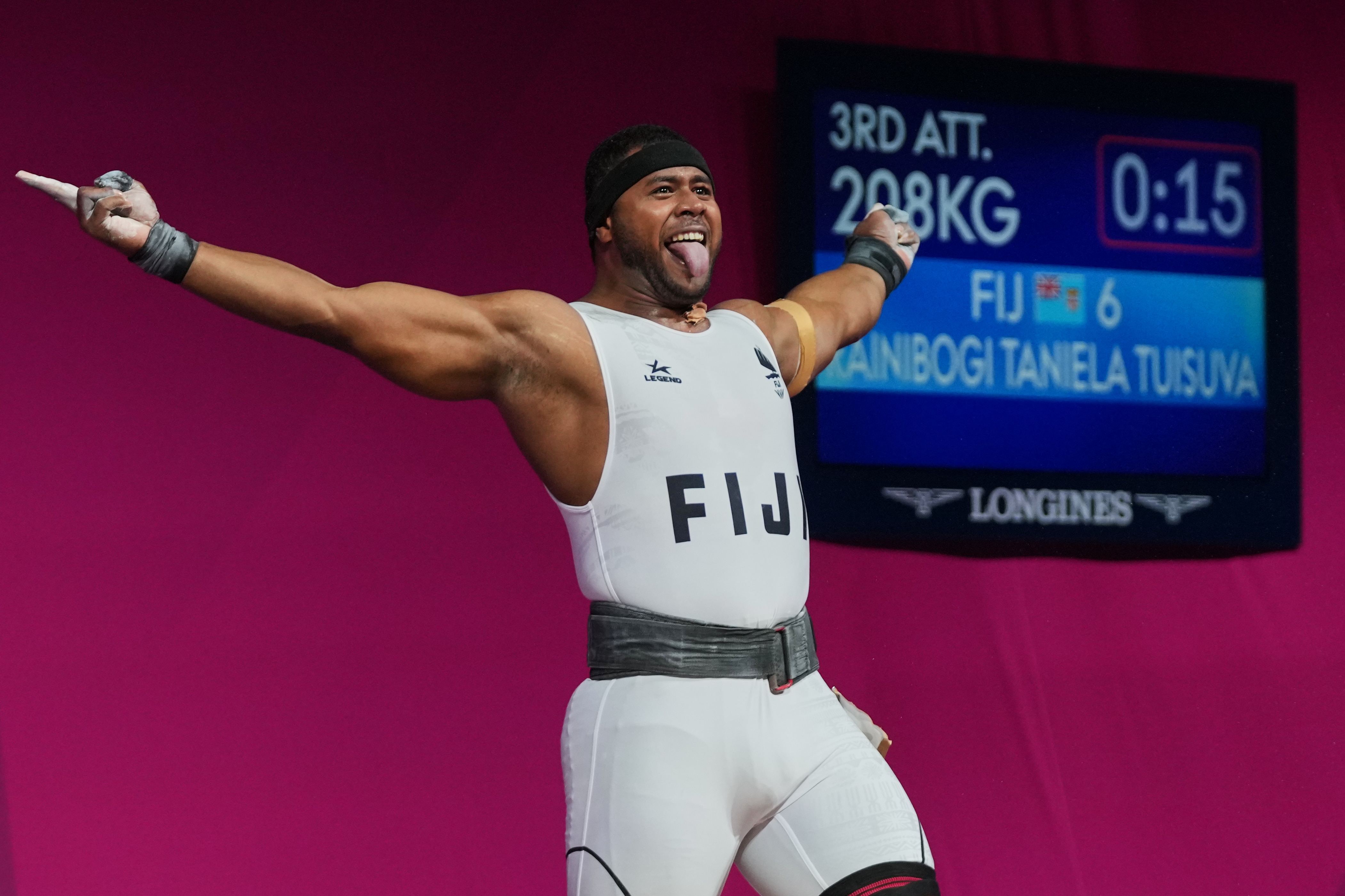 A Fijian man wearing a white weightlifting athletics uniform with FIJI across the front sticks his tongue and arms out