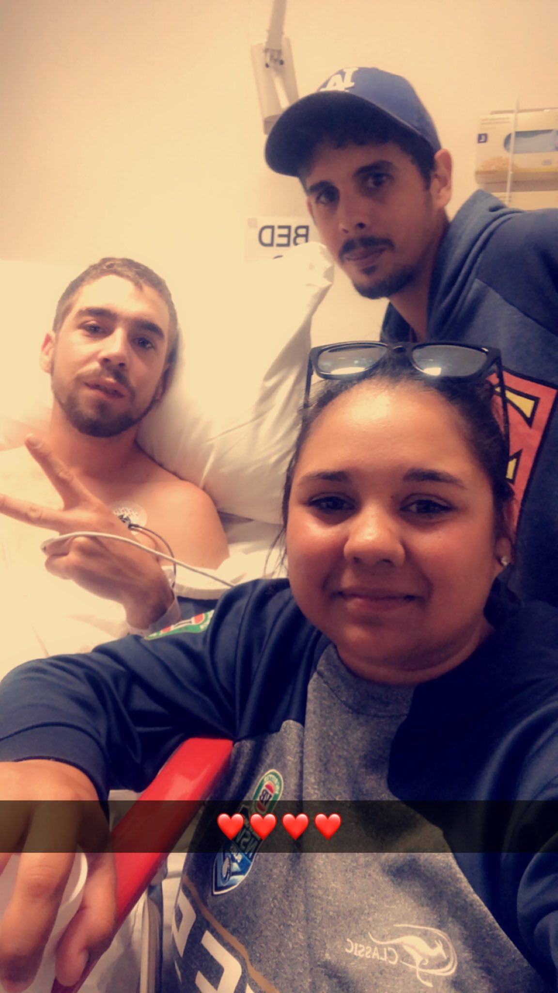 Un hombre con barba está sentado erguido en una cama de hospital, con un hombre y una mujer a su lado sonriendo a la cámara.