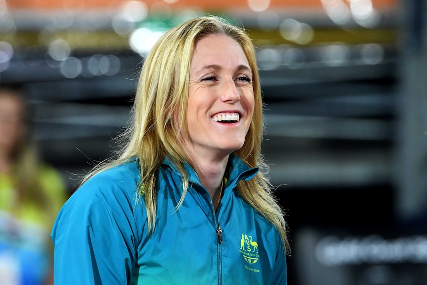 A young blonde white woman wearing a tracksuit top with an Australian emblem smiles