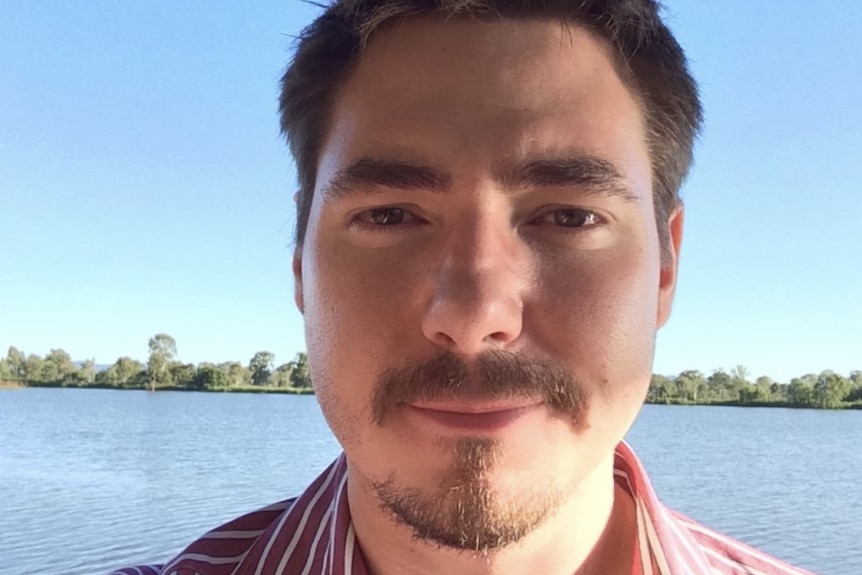 A man with dark hair and a red and white shirt looks at the camera. There is a river in the background.