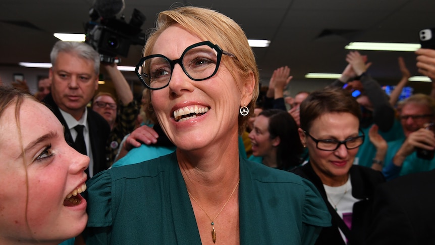 Zoe Daniel grins, surrounded by supporters, all wearing teal, at campaign headquarters.