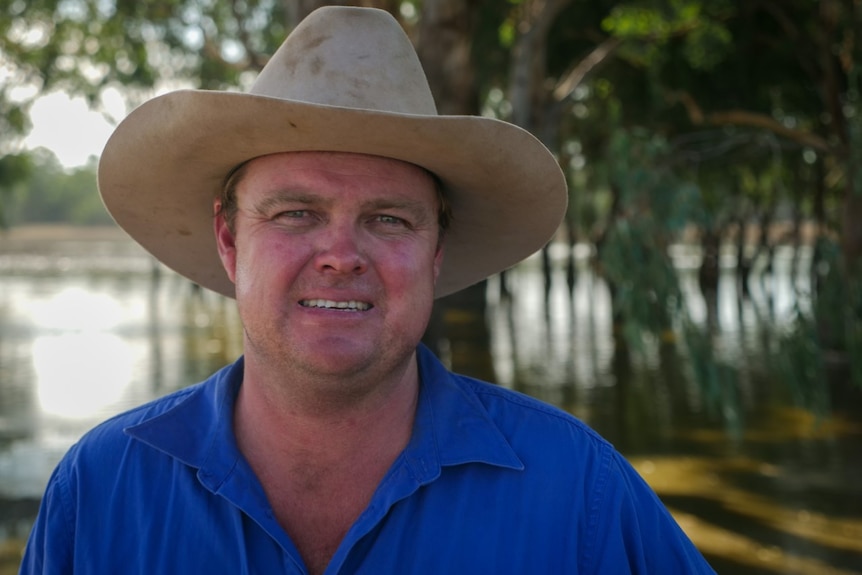 A man in a blue shirt and a hat standing in front of trees and water.