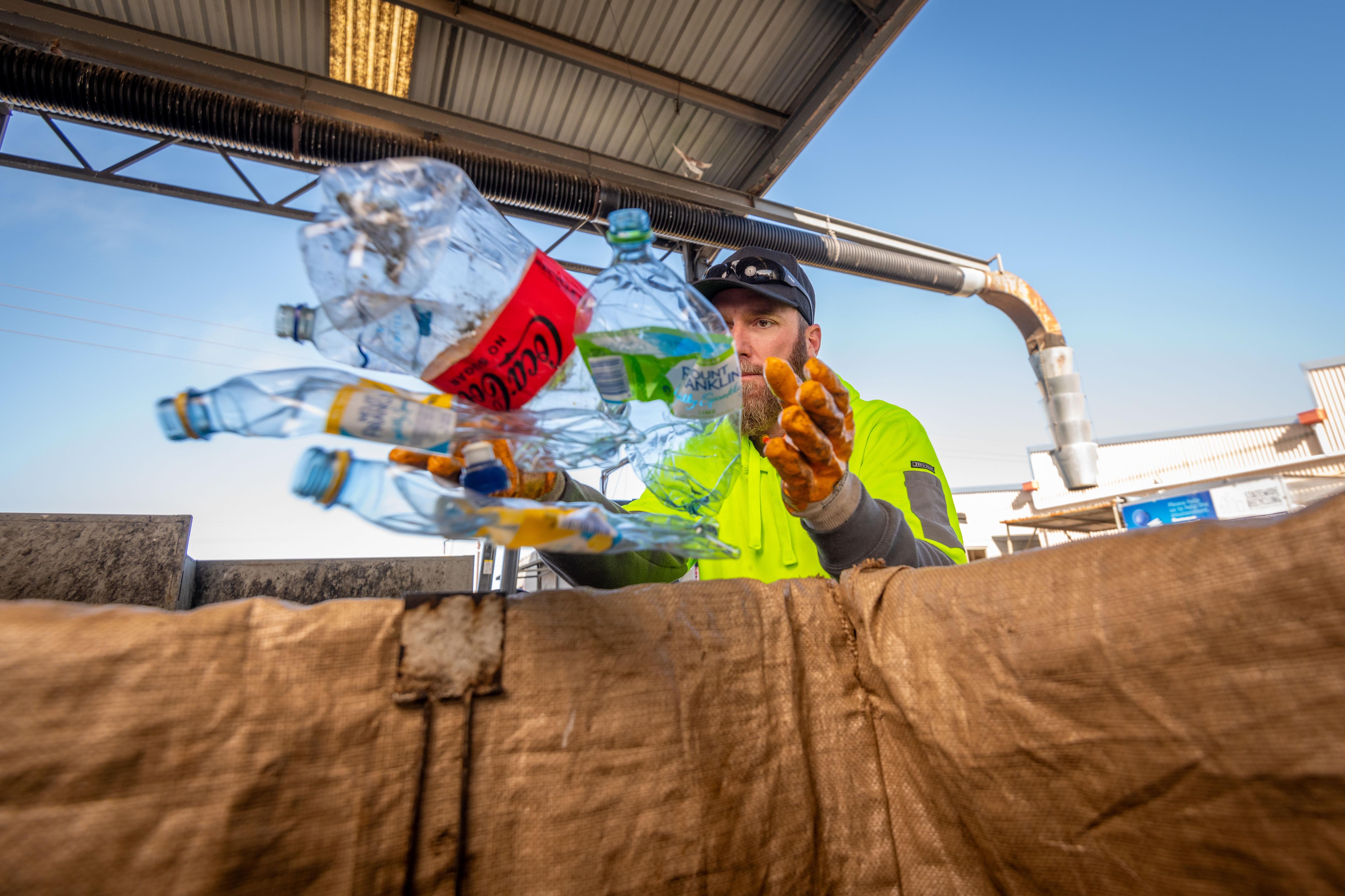 A man wearing a hi-vis hoodie throwing empty bottles into a bin. 
